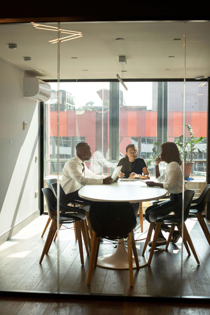 Three office workers sit at a table in a glass conference room